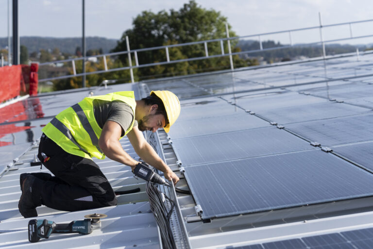 Ein Solateur der Firma IngEne steckt Stromkabel der neuen mit Solarpanels auf dem Dach des Eisfeldes der Winter und Sports World Wallisellen zusammen, fotografiert am Donnerstag, 6. Oktober 2022 in Wallisellen. Die Werke Versorgung Wallisellen AG hat an der Aussenfassade, dem Dach des Eisfeldes sowie der Garderoben und dem Restaurant der Sportanlage eine Gesamtflaeche von insgesamt 3'738 m2 mit Solarpanels verbaut. Diese PV-Anlage produziert bei einer Leistung von 643.4 kWp gesamthaft 650'000 kwh Energie. Ueber das Jahr gerechnet wird die Winter und Sports World Wallisellen damit zum Selbstversorger. Ueberschuesse der erneuerbaren Sonnenenergie kann die Walliseller Bevoelkerung beziehen. (KEYSTONE/Gaetan Bally)