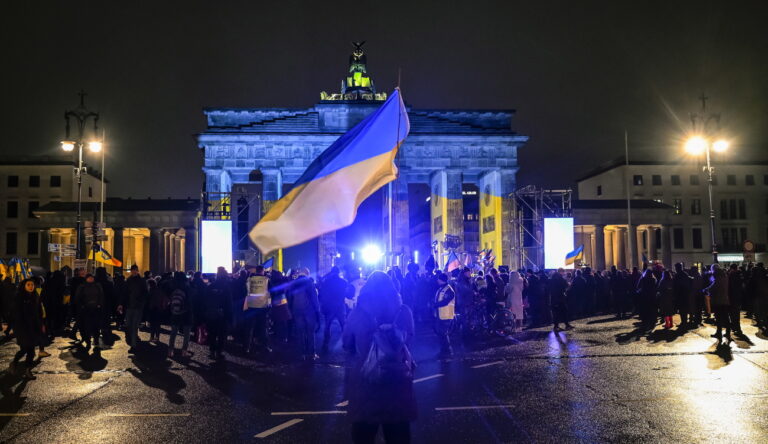 epa10488754 People stand in front of the Brandenburg Gate as it is illuminated in the colors of the Ukrainian flag to mark the first anniversary of the Russian invasion of Ukraine, in Berlin, Germany, 24 February 2023. Russian troops entered Ukrainian territory on 24 February 2022, starting a conflict that has provoked destruction and a humanitarian crisis. One year on, fighting continues in many parts of the country. EPA/FILIP SINGER